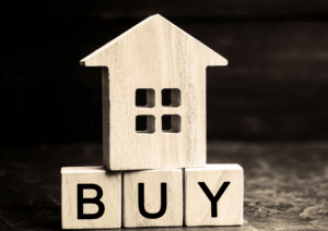 Wooden house model resting on blocks spelling “BUY,” representing the purchase of a rental investment property